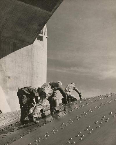 Three construction workers putting a coat of paint on a slanted wall of riveted-steel plates on the Hoover Dam spillway. Photo dated between 1936 and 1946. (United States, Bureau of Reclamation, Library of Congress Prints and Photographs Division Washington, D.C.)